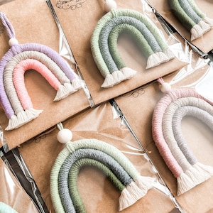 May include: A collection of colourful rainbow wall hangings with white tassels. Each rainbow is made of yarn and has a wooden bead at the top.