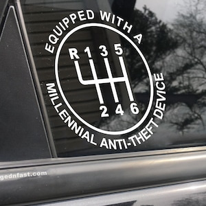 Puede incluir: Calcoman&iacute;a blanca en la ventana de un coche con un patr&oacute;n de cambio de marchas y el texto "EQUIPPED WITH A MILLENNIAL ANTI-THEFT DEVICE". El sitio web "forgednfast.com" est&aacute; impreso debajo. El fondo muestra un reflejo de &aacute;rboles.