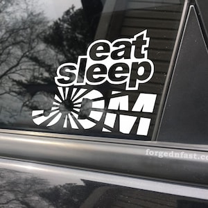 May include: A white vinyl sticker with the text "eat sleep JDM" on a black car window. The JDM text is stylized with a rising sun graphic.