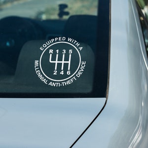 Puede incluir: Una calcoman&iacute;a blanca en la ventana de un coche muestra el patr&oacute;n de cambio de marchas de una transmisi&oacute;n manual. El texto alrededor del patr&oacute;n de cambio dice: "EQUIPADO CON UN DISPOSITIVO ANTI-ROBO MILLENNIAL".