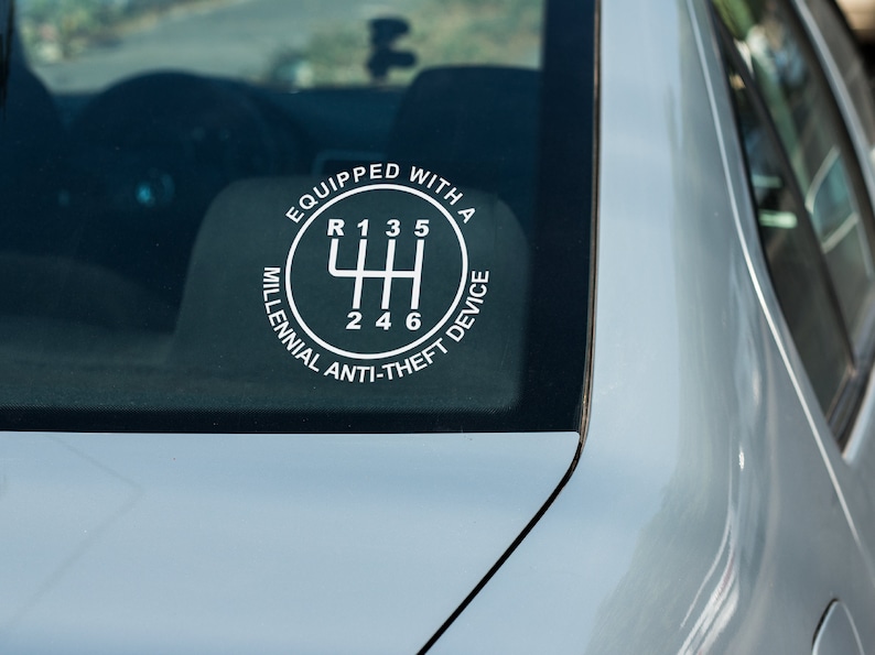 Puede incluir: Una calcoman&iacute;a blanca en la ventana de un coche muestra el patr&oacute;n de cambio de marchas de una transmisi&oacute;n manual. El texto alrededor del patr&oacute;n de cambio dice: "EQUIPADO CON UN DISPOSITIVO ANTI-ROBO MILLENNIAL".