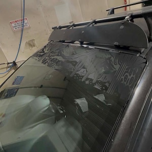 May include: A black car with a decorative desert scene etched on the rear window. A black roof rack and a wind deflector are mounted on the roof. The car is parked inside a garage or car wash.