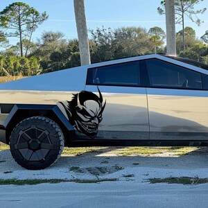 May include: A silver pickup truck with a black and white graphic of a demon on the side. The truck has black wheels and a black trim around the windows.