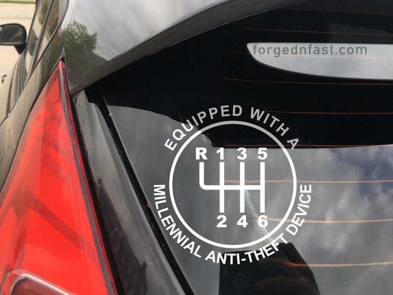 Puede incluir: Calcoman&iacute;a blanca en la ventana de un coche con un diagrama de cambio de marchas y el texto &laquo;EQUIPPED WITH A MILLENNIAL ANTI-THEFT DEVICE&raquo;. Tambi&eacute;n se ve el sitio web &laquo;forgednfast.com&raquo;. El piloto trasero del coche es rojo.