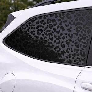 May include: A white vehicle features a black leopard print decal applied to the rear side window. The pattern consists of dark, irregular spots covering the entire glass surface, contrasting against the white body of the car and the red taillight.
