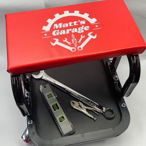 May include: A red rolling mechanic stool with a black frame and a padded seat featuring the white text Matt's Garage, a gear icon, and various tool graphics. A silver 1-1/16 inch wrench, a spirit level, and locking pliers rest on the black lower tray.