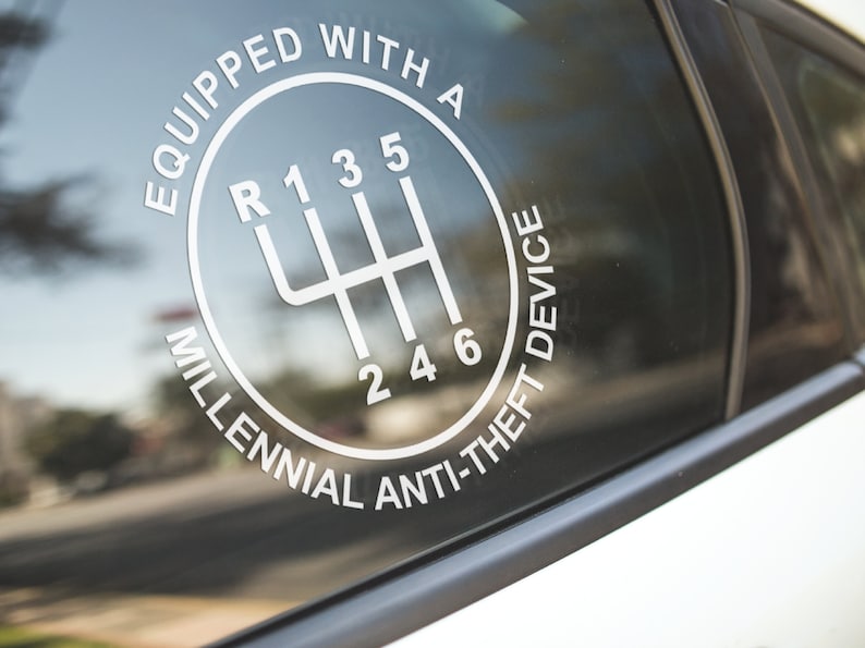 Puede incluir: Calcoman&iacute;a blanca en la ventana de un coche con un patr&oacute;n de cambio de marchas y el texto "EQUIPPED WITH A MILLENNIAL ANTI-THEFT DEVICE". El dise&ntilde;o incluye los n&uacute;meros y posiciones de una transmisi&oacute;n manual de seis velocidades.