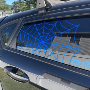 May include: A blue spiderweb decal is applied to the rear window of a black vehicle. The spiderweb design is centered on the window and is a bright blue color. The text "20x2" is visible on the window.