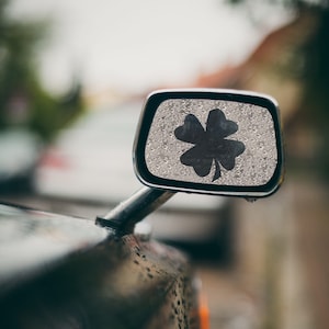 May include: A silver car side mirror with a four-leaf clover sticker on it. The mirror is covered in water droplets.