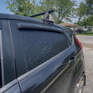 May include: A black car window insert featuring a repeating hexagonal pattern. The insert is installed within the rear passenger side window frame of a black vehicle, positioned below a black roof rack crossbar and a black window rain guard.