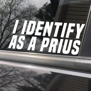 May include: A white vinyl sticker with the text "I IDENTIFY AS A PRIUS" on a black car window.