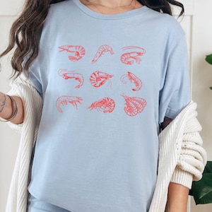May include: A light blue t-shirt with a red shrimp design. The shirt has a crew neck and short sleeves. The shrimp are arranged in a grid pattern on the front of the shirt.