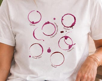 May include: A white short-sleeved t-shirt featuring a graphic print of various red wine glass stains in shades of burgundy and maroon. The design includes labels for wine varieties such as Syrah, Cabernet Sauvignon, Malbec, Riesling, Pinot Noir, and Merlot.