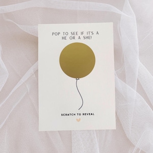 Könnte beinhalten: Eine weiße Karte mit dem Text "POP TO SEE IF IT'S A HE OR A SHE!" über einem goldenen Ballon. Unter dem Ballon steht der Text "SCRATCH TO REVEAL" und ein kleines rosa Herz.