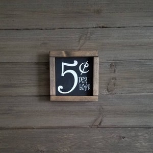 May include: A small wooden sign with a black chalkboard insert. The chalkboard has white text that reads "5&cent; per load".