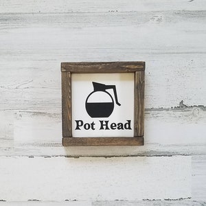 Puede incluir: Un letrero de madera con fondo blanco y texto negro que dice "Pot Head". El letrero presenta una silueta negra de una cafetera.