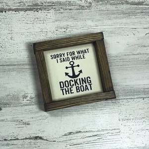Op de afbeelding: Een houten ingelijst bord met een witte achtergrond en zwarte tekst die luidt "Sorry voor wat ik zei tijdens het aanmeren van de boot". Een ankersymbool bevindt zich boven de tekst.