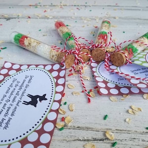 Puede incluir: Un conjunto de cinco tubos de ensayo de vidrio llenos de una mezcla de avena, chispas y azúcar. Los tubos de ensayo están atados con un cordel rojo y blanco y tienen tapones de corcho. Los tubos de ensayo están sobre una superficie de madera blanca con una tarjeta de lunares rojos y blancos que dice "Sprinkle this reindeer food outside tonight. The moonlight will make it sparkle bright. As the reindeer fly and roam. This will guide them to your home."