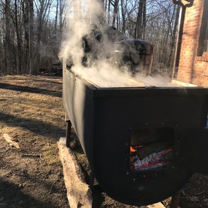Puede incluir: Un evaporador de metal negro con un hogar y una olla grande en la parte superior. El evaporador est&aacute; sentado sobre troncos de madera y est&aacute; rodeado de &aacute;rboles. El vapor se eleva de la olla.
