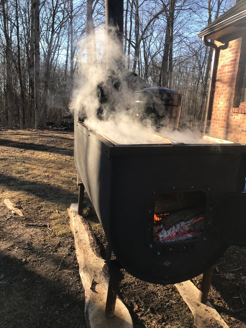 Puede incluir: Un evaporador de metal negro con un hogar y una olla grande en la parte superior. El evaporador est&aacute; sentado sobre troncos de madera y est&aacute; rodeado de &aacute;rboles. El vapor se eleva de la olla.