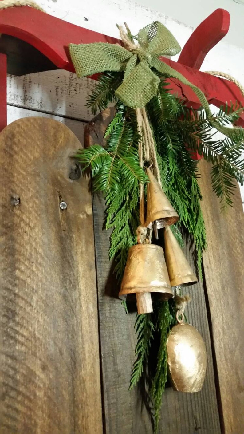 May include: A rustic Christmas decoration featuring a red wooden sled with a green burlap bow and evergreen branches. Gold-colored bells hang from the branches, creating a festive holiday display.