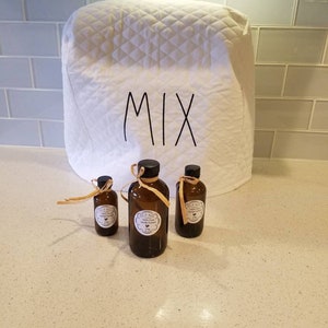 May include: Three glass bottles with brown liquid and labels that say "100% Pure Vanilla Extract" on a white countertop. The bottles are in front of a white quilted kitchen appliance cover with the word "MIX" written on it.