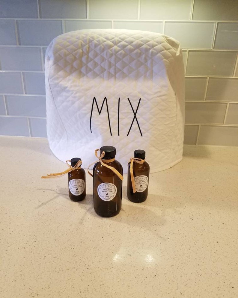 May include: Three glass bottles with brown liquid and labels that say "100% Pure Vanilla Extract" on a white countertop. The bottles are in front of a white quilted kitchen appliance cover with the word "MIX" written on it.