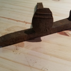 May include: A rusty, antique railroad spike, featuring a flat head with stamped markings and a tapered, pointed end. The spike is made of iron and rests on a light-colored wooden surface. The overall design suggests a tool used in railway construction.