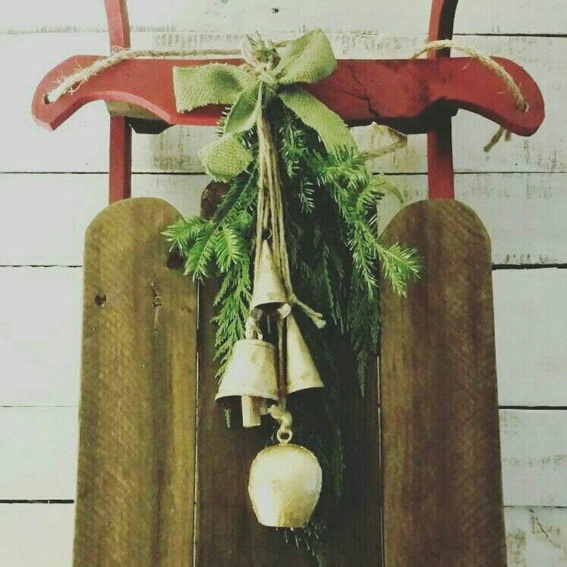 May include: A rustic wooden sled with a red top and wooden runners. The sled is decorated with evergreen branches, a burlap bow, and three gold-colored bells, creating a festive holiday decoration.