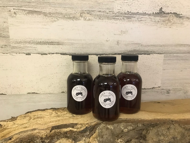 Puede incluir: Tres botellas de vidrio de jarabe de arce con tapas negras y etiquetas blancas que muestran un dise&ntilde;o de tractor. Las etiquetas tambi&eacute;n incluyen el texto "Cinnamon Hill Farm" y "Jarabe de arce puro". Las botellas est&aacute;n sobre una superficie de madera.