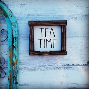 Könnte beinhalten: Ein Holzschild mit weißem Hintergrund und schwarzem Text, der "TEA TIME" lautet. Das Schild ist in dunkelbraunem Holz gerahmt und hat einen rustikalen Look.