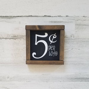 May include: A wooden sign with a black background and white lettering. The sign reads "5&cent; per load".