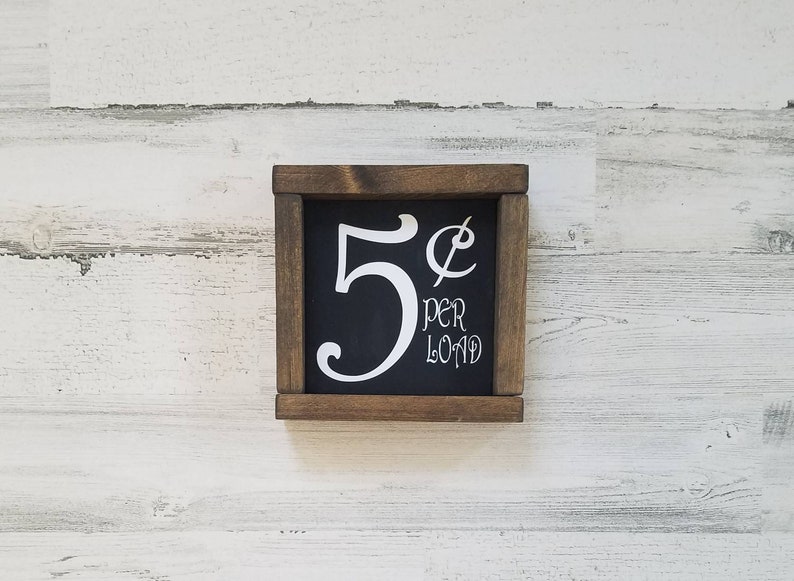 May include: A wooden sign with a black background and white lettering. The sign reads "5&cent; per load".
