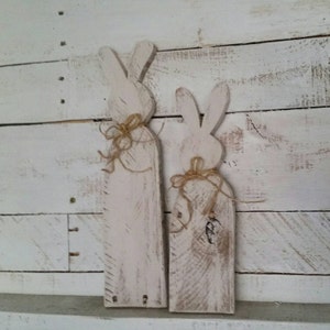 May include: Two white wooden bunny shapes with a rustic finish. The bunnies are tied with twine bows and stand upright on a white wood background.