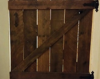 Könnte beinhalten: Eine rustikale Holztür mit einer diagonalen Strebe und schwarzen Metallscharnieren. Die Tür ist aus wiederverwendeten Holzbrettern gefertigt und hat eine dunkelbraune Lasur.