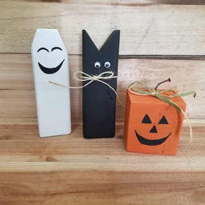 Peut inclure: Trois décorations d'Halloween en bois : un fantôme blanc avec un visage souriant, un chat noir avec des yeux globuleux et une citrouille orange avec un visage de citrouille.