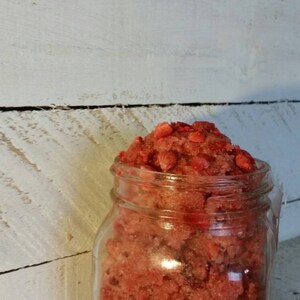 May include: A glass jar filled with a red and white sugar scrub. The scrub is made with sugar and fresh strawberries.