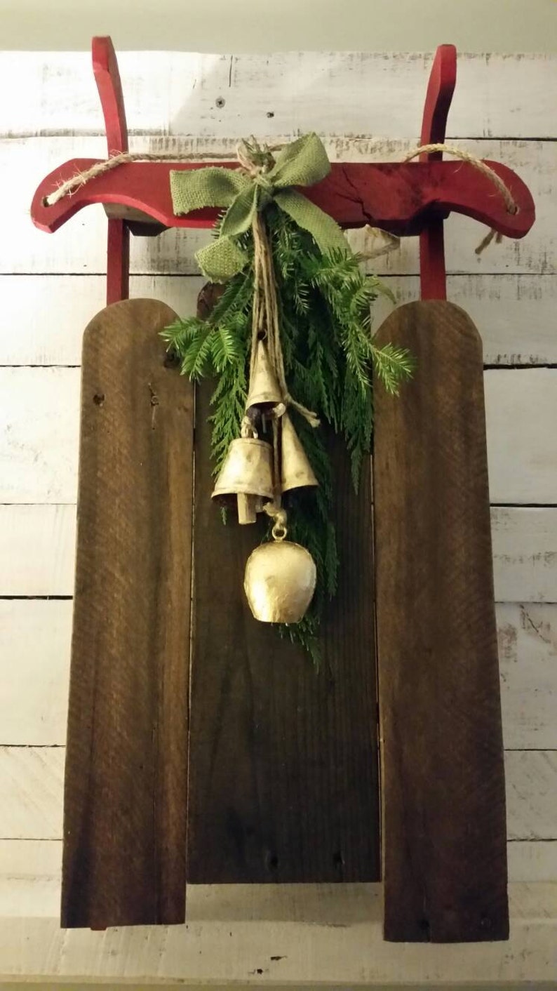 May include: A rustic wooden sled with a red frame, adorned with a green bow, evergreen branches, and gold bells. The sled is set against a white, weathered wood background, creating a charming, festive display.