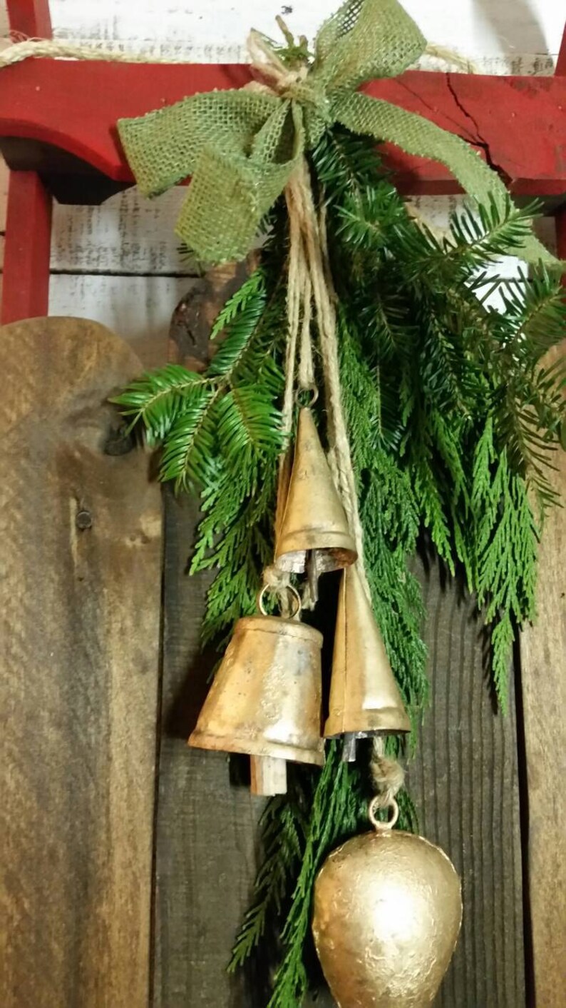 May include: A rustic Christmas decoration featuring three gold-colored bells of varying sizes, suspended from twine and evergreen branches. A burlap bow tops the arrangement, set against a wooden background.