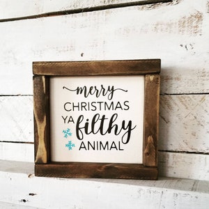 May include: A wooden sign with a white background and brown frame. The sign says "merry christmas ya filthy animal" with two blue snowflakes.