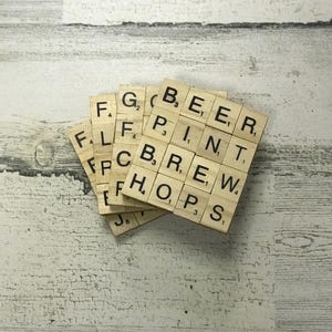 May include: Four square wooden coasters made from Scrabble tiles. The coasters spell out "BEER PINT BREW HOPS". The tiles are light brown with black letters and numbers. The coasters are arranged on a distressed white wooden surface.