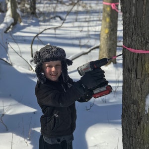Puede incluir: Una persona joven con una chaqueta negra y un gorro de piel perfora un agujero en el tronco de un &aacute;rbol en un bosque nevado. La persona est&aacute; usando un taladro inal&aacute;mbrico con un mango rojo y negro. Hay una cinta rosa atada alrededor del tronco del &aacute;rbol.