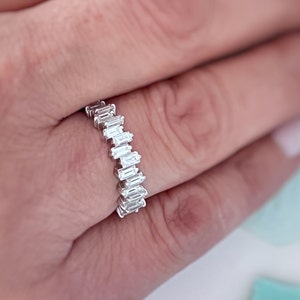 Peut inclure: Une bague en argent ornée d'une série de pierres précieuses rectangulaires et transparentes. La bague est portée au doigt d'une personne, avec la main et une partie de l'arrière-plan visibles. Les pierres sont disposées en quinconce.