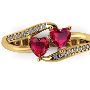 Puede incluir: Un anillo de oro con dos piedras preciosas rojas en forma de corazón. La banda es curva y está adornada con pequeñas piedras preciosas transparentes. El anillo se muestra sobre un fondo blanco, resaltando el diseño y el brillo de la joya.