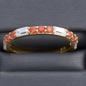 May include: A gold band ring with alternating orange and clear gemstones. The ring is set in a simple band.
