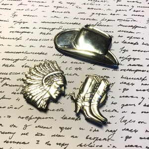 Peut inclure: Trois épingles en métal argenté représentant un chapeau de cowboy, une botte de cowboy et une coiffe d'Indien d'Amérique. Les épingles sont sur un fond blanc avec du texte manuscrit.