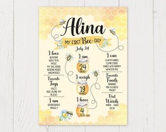 May include: A yellow and white bee-themed first birthday chart featuring the name Alina. The chart includes details about the child's age, height, weight, favourite toys, foods, and first words. The chart is decorated with bees, flowers, and honeycomb patterns.