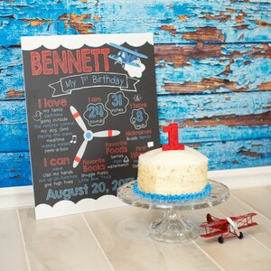 May include: A chalkboard sign with the name Bennett and details about his first birthday. The sign includes his weight, height, favorite foods, books, and activities. A small white cake with a red number one candle sits on a glass cake stand. A red toy airplane is on the floor in front of the cake.