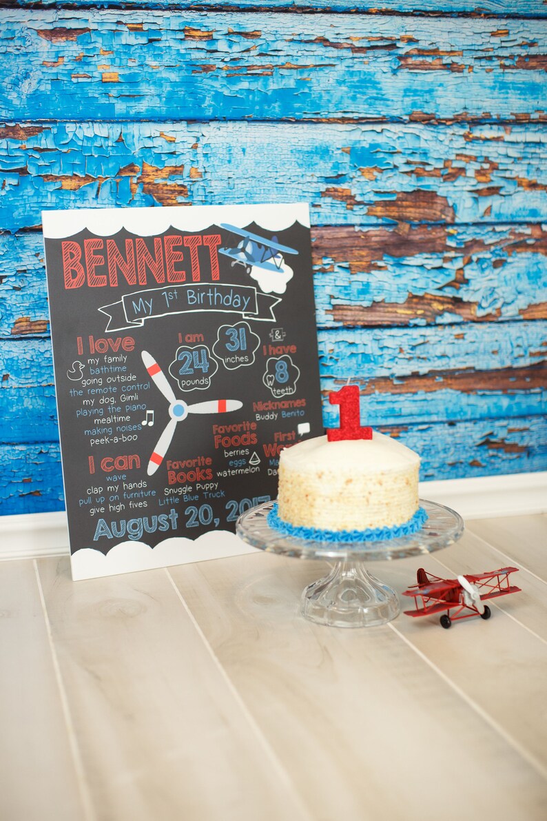 May include: A chalkboard sign with the name Bennett and details about his first birthday. The sign includes his weight, height, favorite foods, books, and activities. A small white cake with a red number one candle sits on a glass cake stand. A red toy airplane is on the floor in front of the cake.