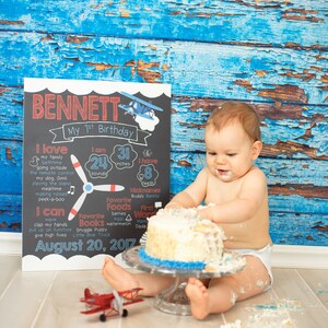 May include: A baby boy sits on a wooden floor with a blue and white cake in front of him. He is wearing a white diaper and has frosting on his face and hands. A chalkboard sign behind him says "Bennett My 1st Birthday" and lists his age, weight, height, and other details. A red toy airplane is on the floor in front of him.